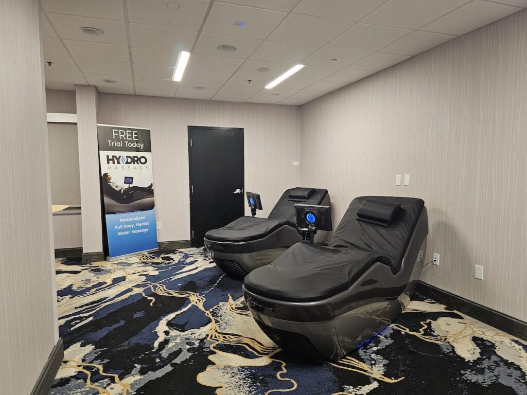 A room with two HydroMassage beds covered in black leather, positioned side by side on a patterned carpet. A sign next to the beds offers a free trial of the HydroMassage, describing it as a personalized, full-body, heated water massage. The room has light-colored walls and a closed black door in the background.