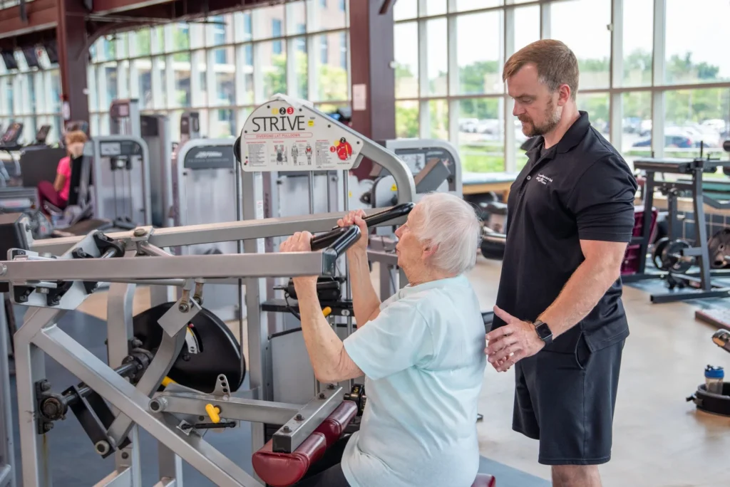 Older woman using a weight machine with help from a male trainer in a gym.