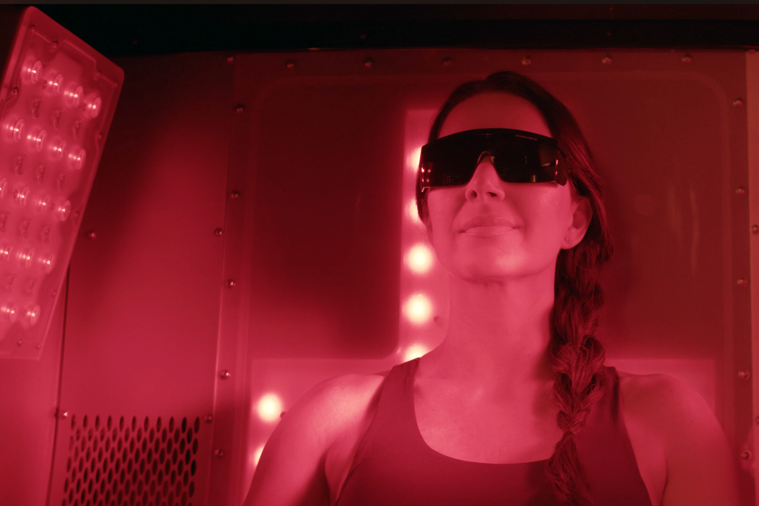 Product - RedZone Sauna 2 Woman wearing dark protective glasses sits inside the red-lit sauna chamber with LED panels glowing beside her.