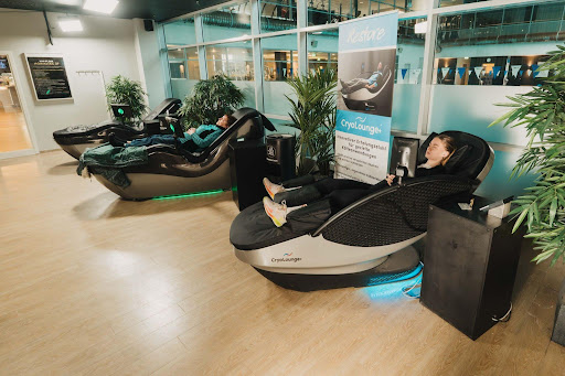 Two people relaxing in reclining CryoLounge massage chairs in a bright wellness lounge, surrounded by plants and soft lighting.